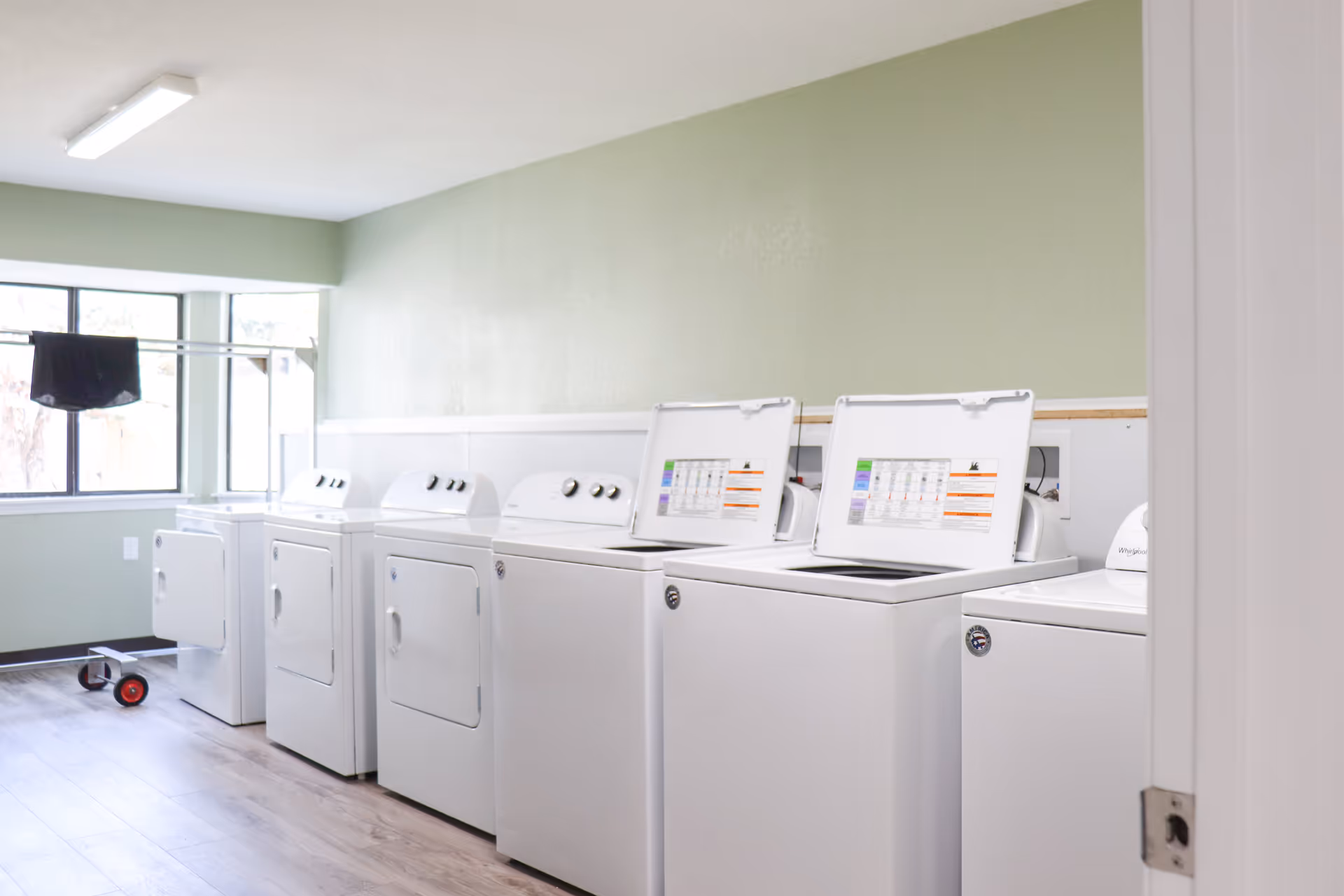 Laundry room with a row of white washing machines and dryers against a pale green wall and a window.