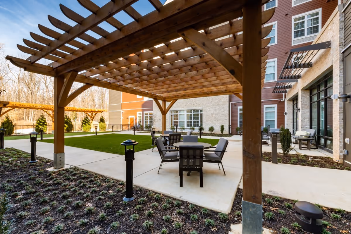 Outdoor seating area at Tribute at Black Hill featuring a wooden pergola over a concrete patio with tables and chairs. The area is surrounded by landscaped plants, a green lawn, and the exterior of a multi-story building with large windows.