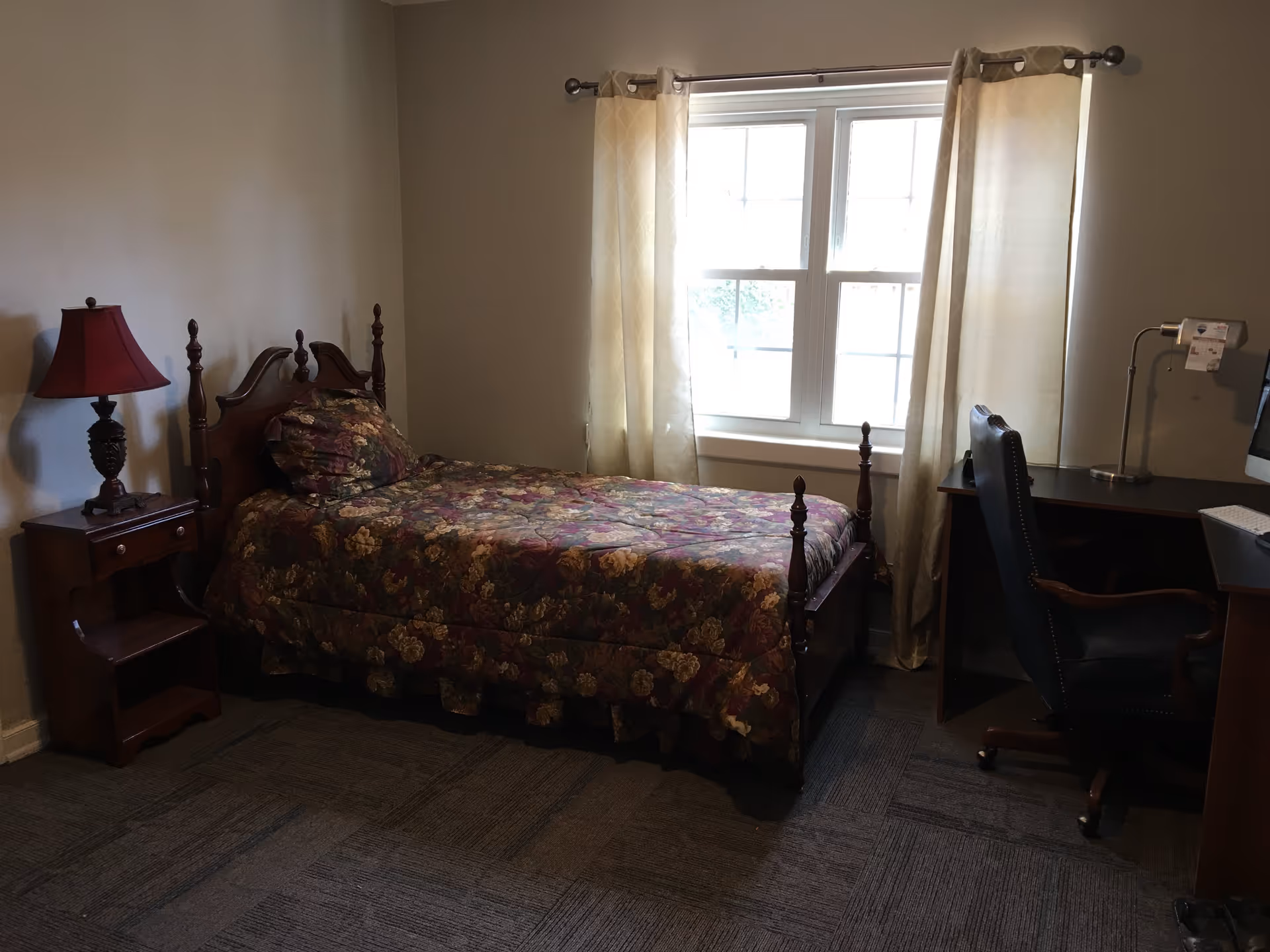 Bedroom with a single floral bed, wooden nightstand and lamp, and a desk and chair beside a curtained window.