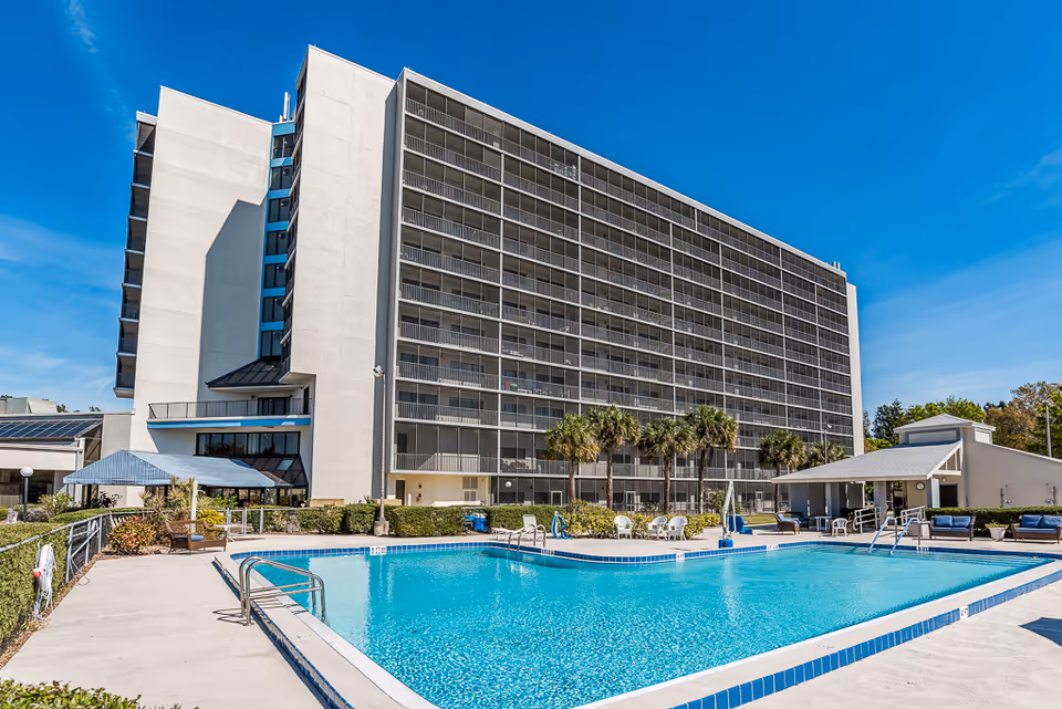 Outdoor view of a multi-story senior living facility building with screened balconies, a clear blue swimming pool in the foreground, palm trees, and seating areas under a sunny blue sky.