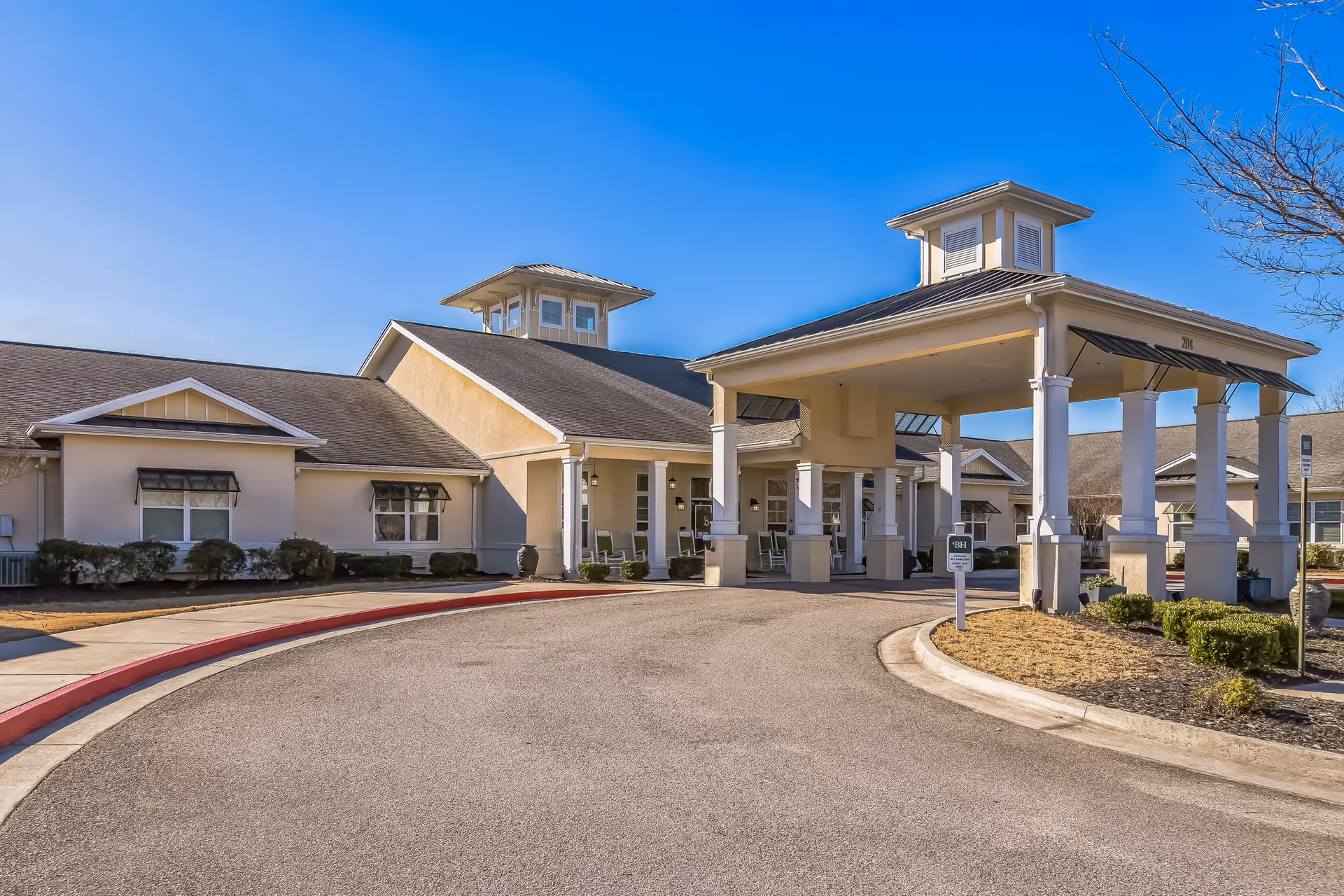 Exterior view of Barclay House of Augusta, showing a single-story building with a covered entrance supported by white columns, surrounded by a curved driveway and landscaped bushes under a clear blue sky.