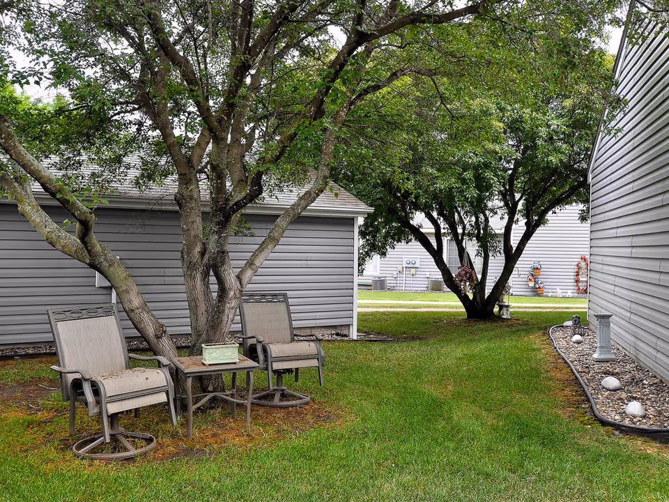 A peaceful outdoor area between buildings with green grass, two metal chairs with cushions, a small table with a planter, and several trees providing shade. The buildings have gray siding and there are decorative rocks along the building edges.
