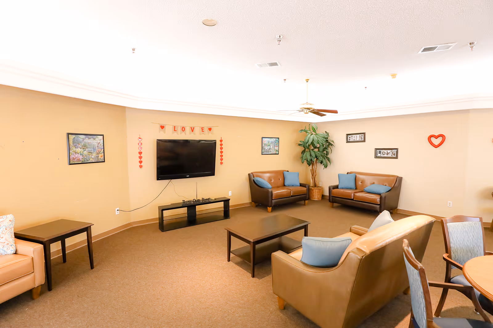 Well-lit communal lounge with sofas and chairs arranged around a coffee table facing a wall-mounted TV and decorative wall art.