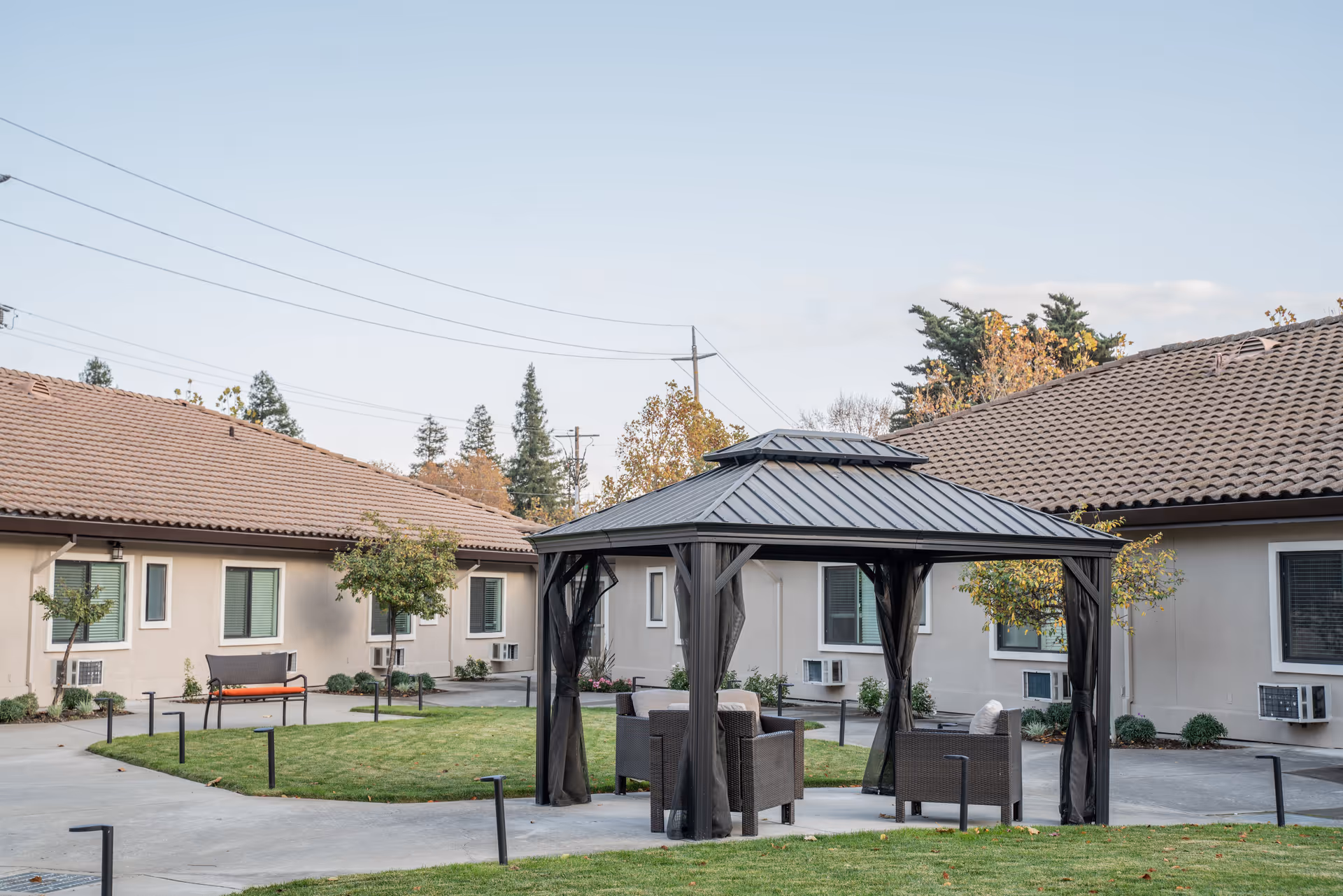 Outdoor courtyard area at Carefield Pleasanton Memory Care featuring a gazebo with seating underneath, surrounded by single-story buildings with tiled roofs, small trees, and a well-maintained lawn.