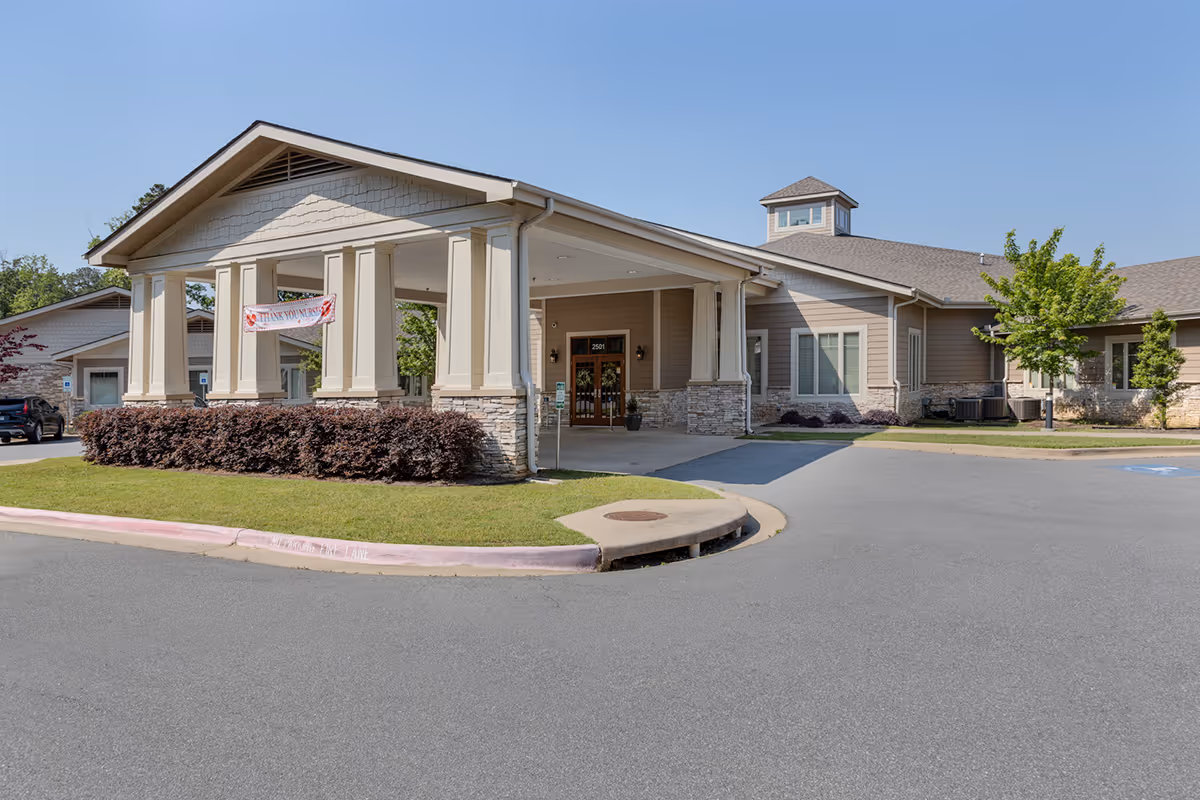 Exterior view of Chapters Living of Little Rock, showing the main entrance with a covered drop-off area supported by columns, beige siding with stone accents, a small tower on the roof, and a clear blue sky.