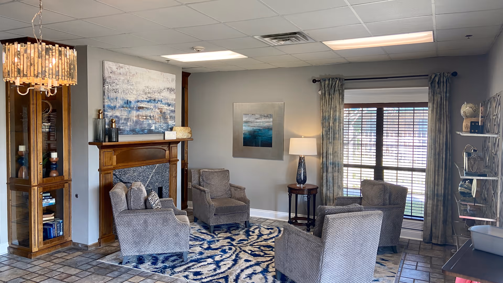 A cozy living room area with four gray upholstered armchairs arranged around a blue and white patterned rug. There is a wooden fireplace with decorative items and a large abstract painting above it. A tall wooden cabinet with glass doors stands to the left, and a window with curtains and blinds is on the right. A small round side table with a lamp is positioned between two chairs near the window. The room has tiled flooring and a ceiling with fluorescent lighting.