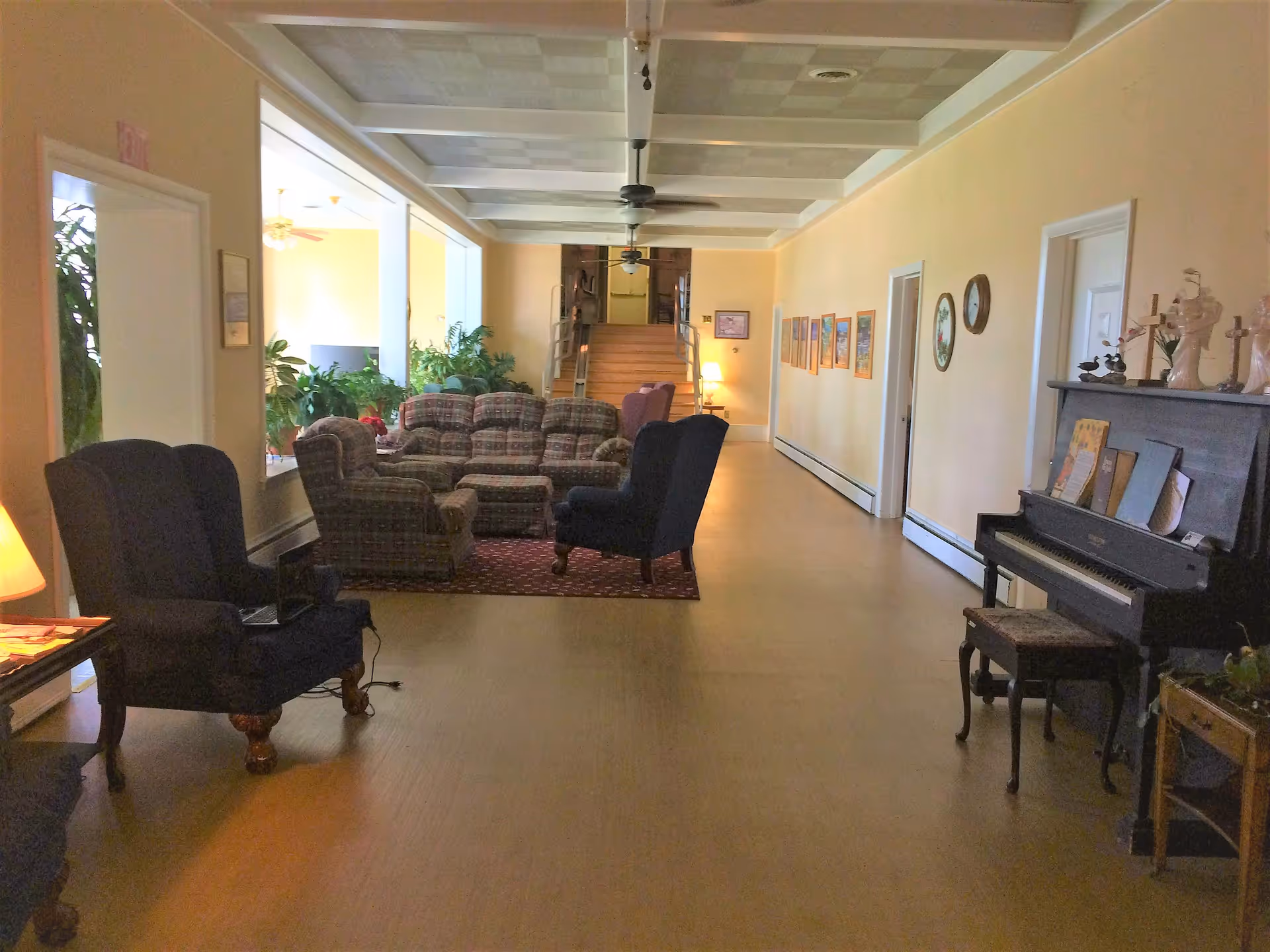 A long, narrow living room area with a mix of plaid and dark upholstered armchairs and sofas arranged on a small rug. There is a piano with a stool on the right side, decorated with figurines and sheet music. The walls are light yellow with framed pictures and a clock. Ceiling fans hang from the coffered ceiling, and there are plants near the large windows on the left side. A staircase is visible at the far end of the room.