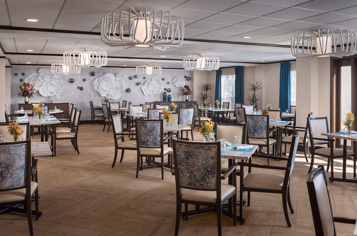 A spacious dining room with multiple tables and chairs arranged neatly. Each table has a small floral centerpiece and place settings with napkins and glasses. The room features modern light fixtures hanging from the ceiling, large windows with blue curtains allowing natural light, and decorative wall art with white floral designs on a gray wall.