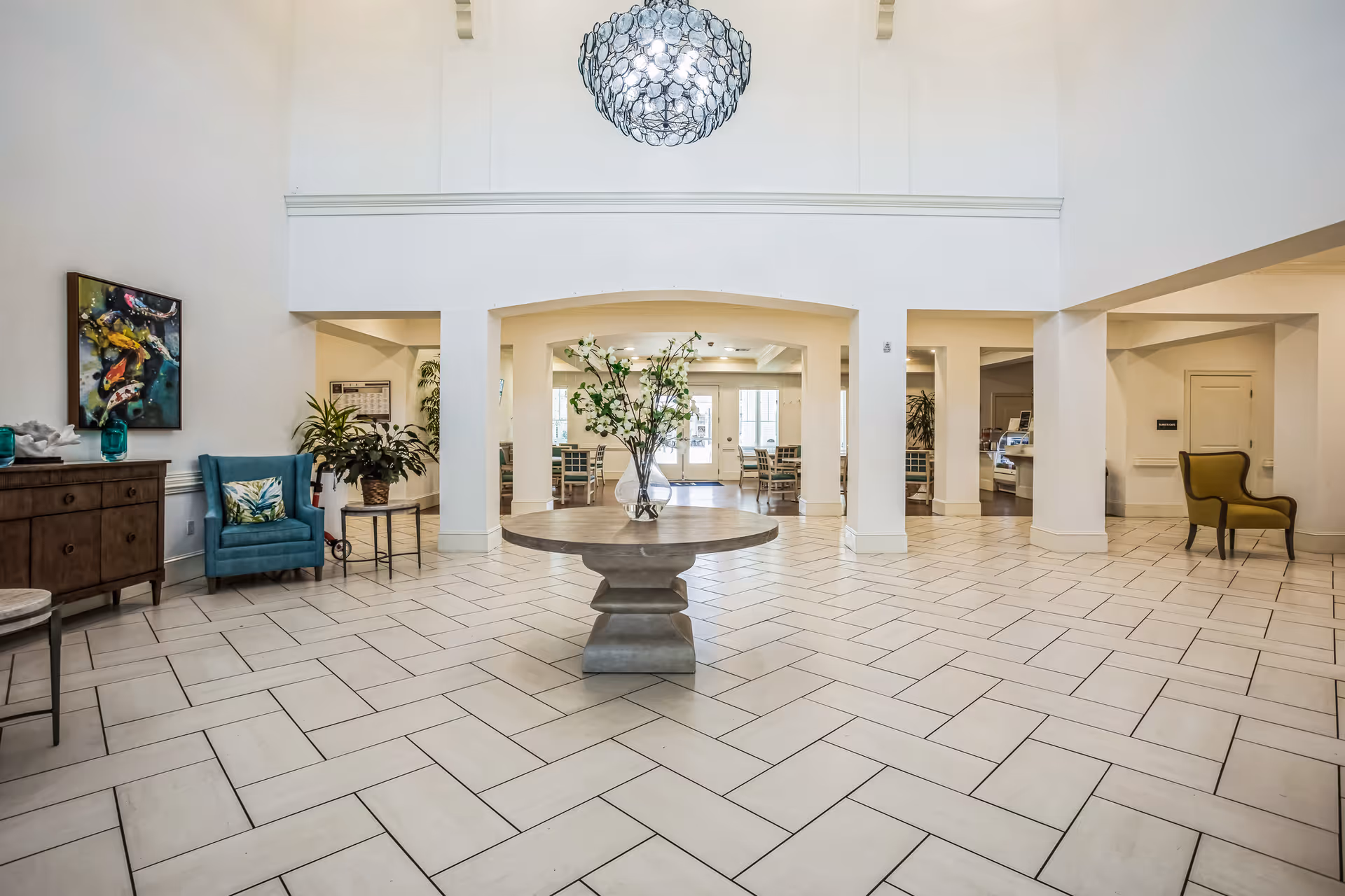 Spacious and bright interior lobby area with a large round table in the center holding a vase with white flowers. The floor is tiled in a herringbone pattern. There are several seating areas with chairs, a wooden sideboard with decorative items, and a large modern chandelier hanging from the ceiling. The walls are painted white, and there is a colorful painting on the left wall.