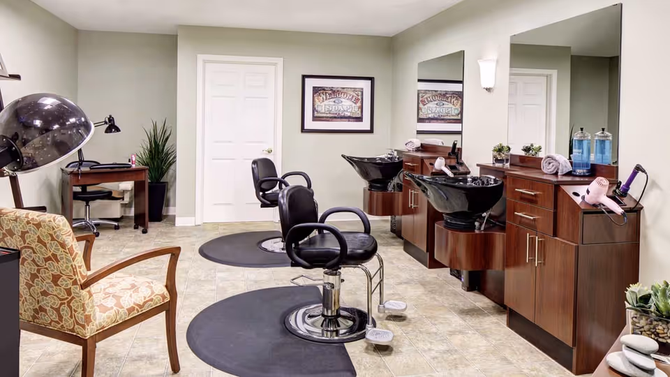 Interior of a hair salon area in a senior living facility featuring two black salon chairs in front of two hair washing stations with large mirrors above wooden cabinets. There is a patterned armchair on the left, a hair dryer hood, a small desk with a lamp and chair in the background, and some decorative plants. The walls are light-colored and the floor is tiled.