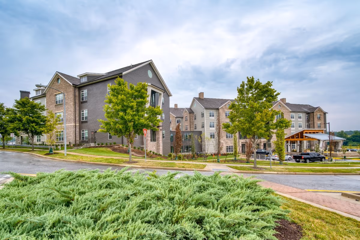 Exterior view of a multi-story senior living facility building with a mix of brick and siding facade, surrounded by green trees and landscaped bushes under a cloudy sky. A driveway and parked vehicles are visible near the entrance.