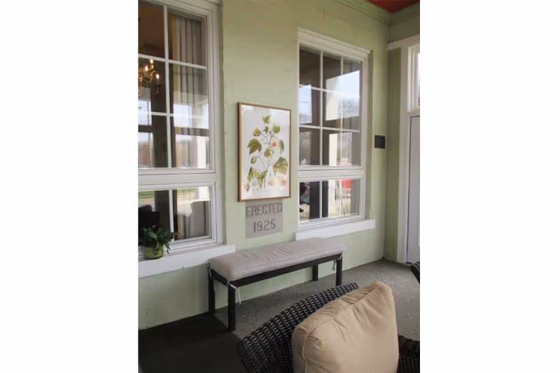 Covered front porch area with a bench beneath windows, botanical wall art, an 'ERECTED 1925' plaque, and wicker seating with cushions.
