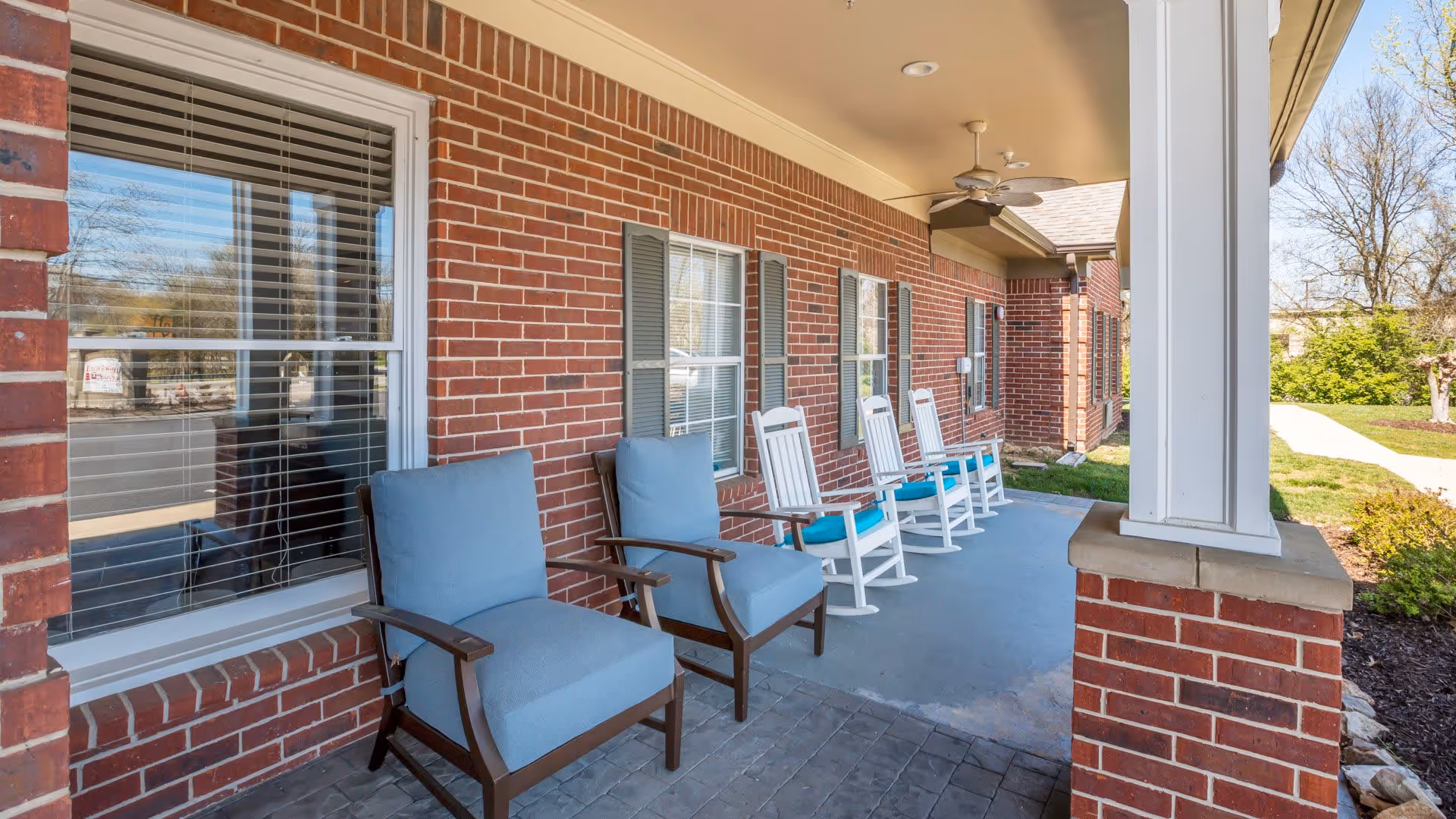 A covered outdoor porch area with brick walls and several chairs, including two cushioned armchairs and four white rocking chairs with blue cushions, overlooking a walkway and landscaped greenery.