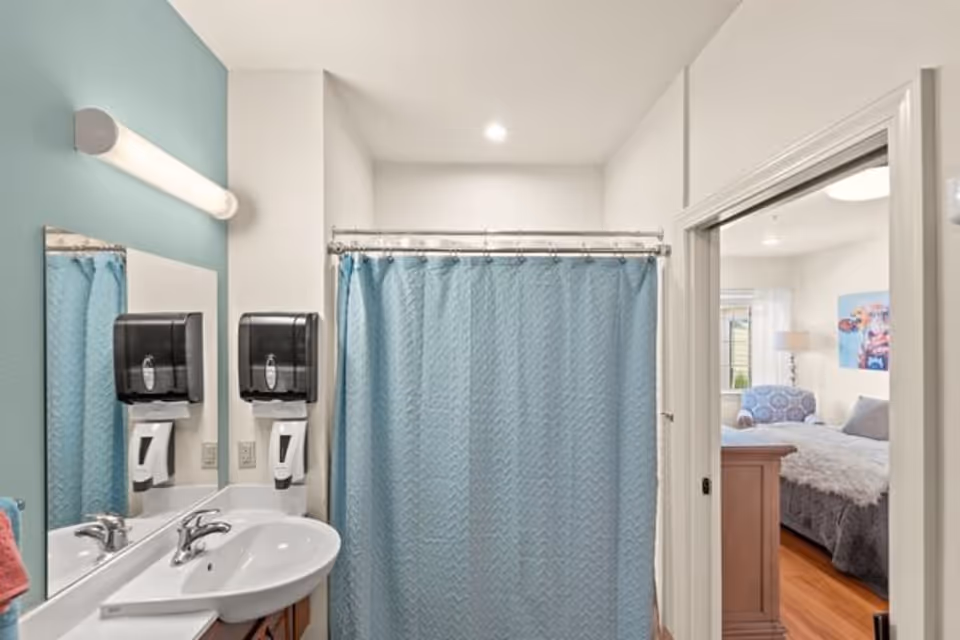 Small bathroom with a sink, mirror, wall-mounted dispensers and a blue shower curtain, with a bedroom visible through the open door.