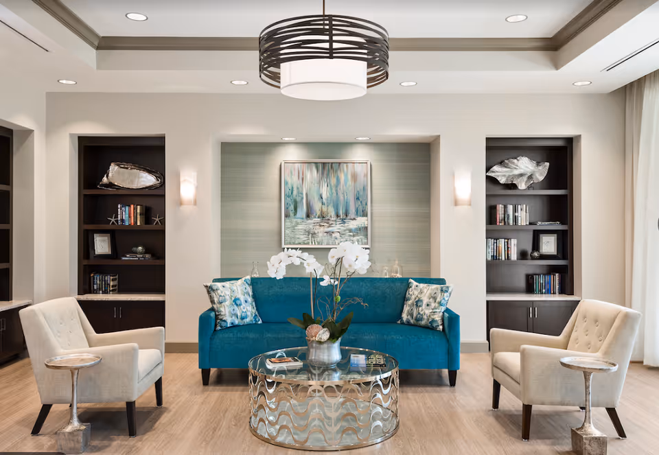 A modern living room with a teal sofa adorned with floral cushions, two beige armchairs with small side tables, a round glass coffee table with a decorative plant, built-in dark wood shelves with books and decor, and a large abstract painting on the wall under soft ceiling lights.