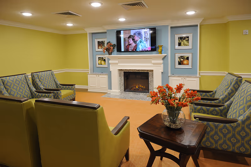 A cozy senior living room with green and patterned armchairs arranged around a small wooden table holding a vase of orange flowers. A white fireplace with a granite surround is centered on the far wall, above which a flat-screen TV is mounted showing a scene from a TV show. The walls are painted light green with a blue accent wall behind the fireplace, decorated with framed fruit artwork.