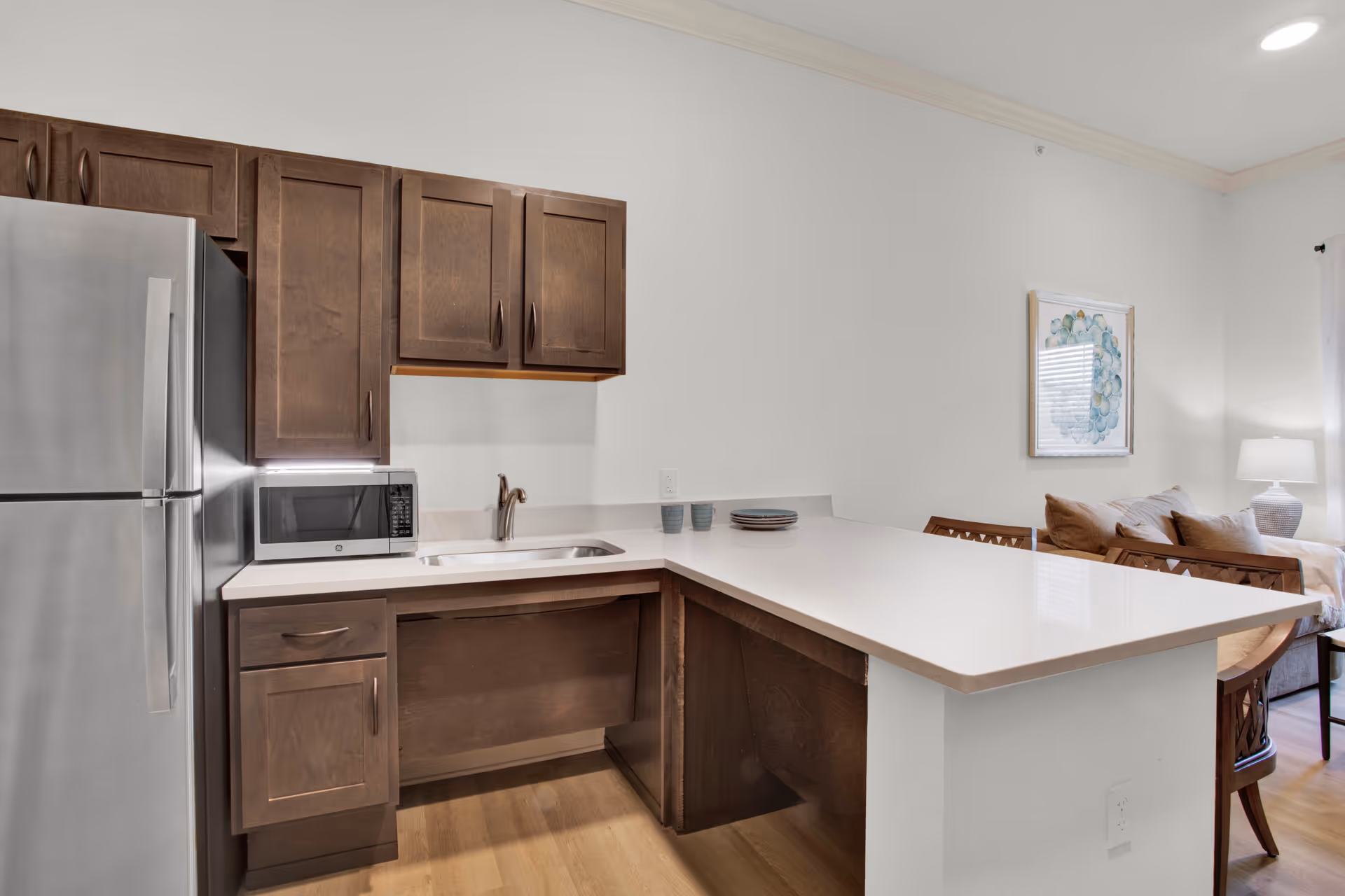 Modern kitchen area with wooden cabinets, a stainless steel refrigerator, a microwave on the counter, a sink, and a white countertop extending into a breakfast bar with two chairs. In the background, part of a living room with a couch, pillows, a lamp, and a framed artwork on the wall is visible.