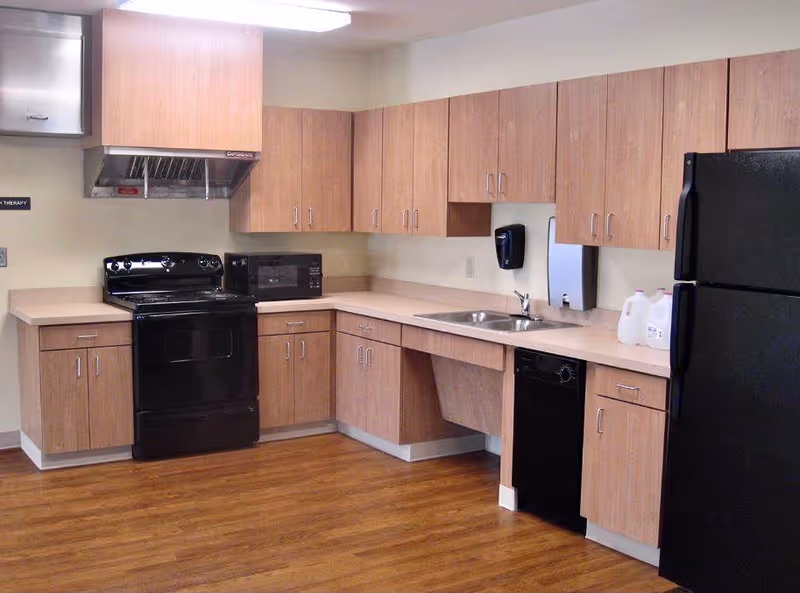 A clean institutional kitchen with wood cabinets, a black stove and refrigerator, microwave, sink, dishwasher, and wood-look flooring.