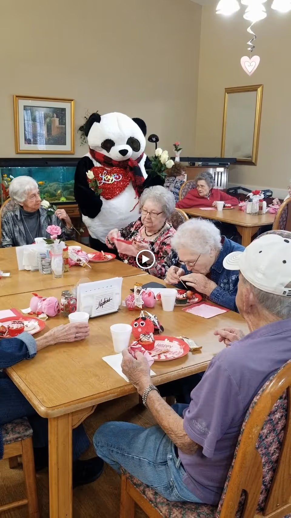 A group of elderly people sitting around a wooden table in a senior living facility dining area, enjoying cupcakes and drinks. A person dressed in a panda costume with a red heart-shaped 'Love' decoration stands behind them. The room has beige walls, framed pictures, and a fish tank in the background.