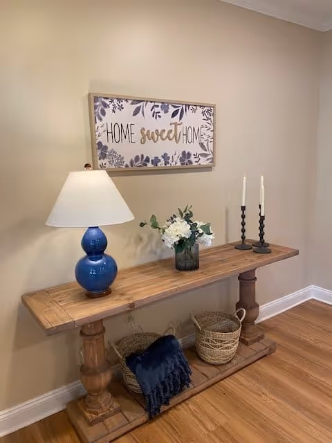 A wooden console table against a beige wall with a blue lamp, a vase of white flowers, and two black candle holders with white candles. Above the table is a framed sign that reads 'Home sweet Home' with floral decorations. Below the table are two woven baskets, one containing a dark blue throw blanket. The floor is wooden.