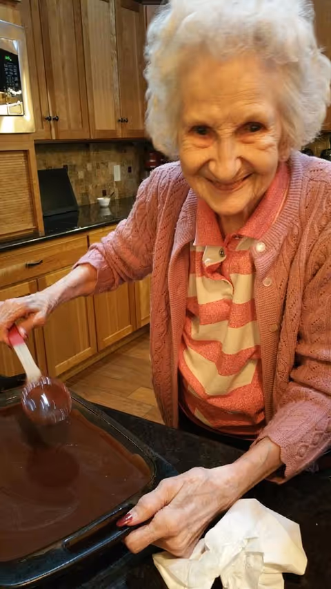 An elderly woman with white hair wearing a pink striped shirt and a pink cardigan is smiling while spreading chocolate batter in a baking pan in a kitchen with wooden cabinets and a black countertop.