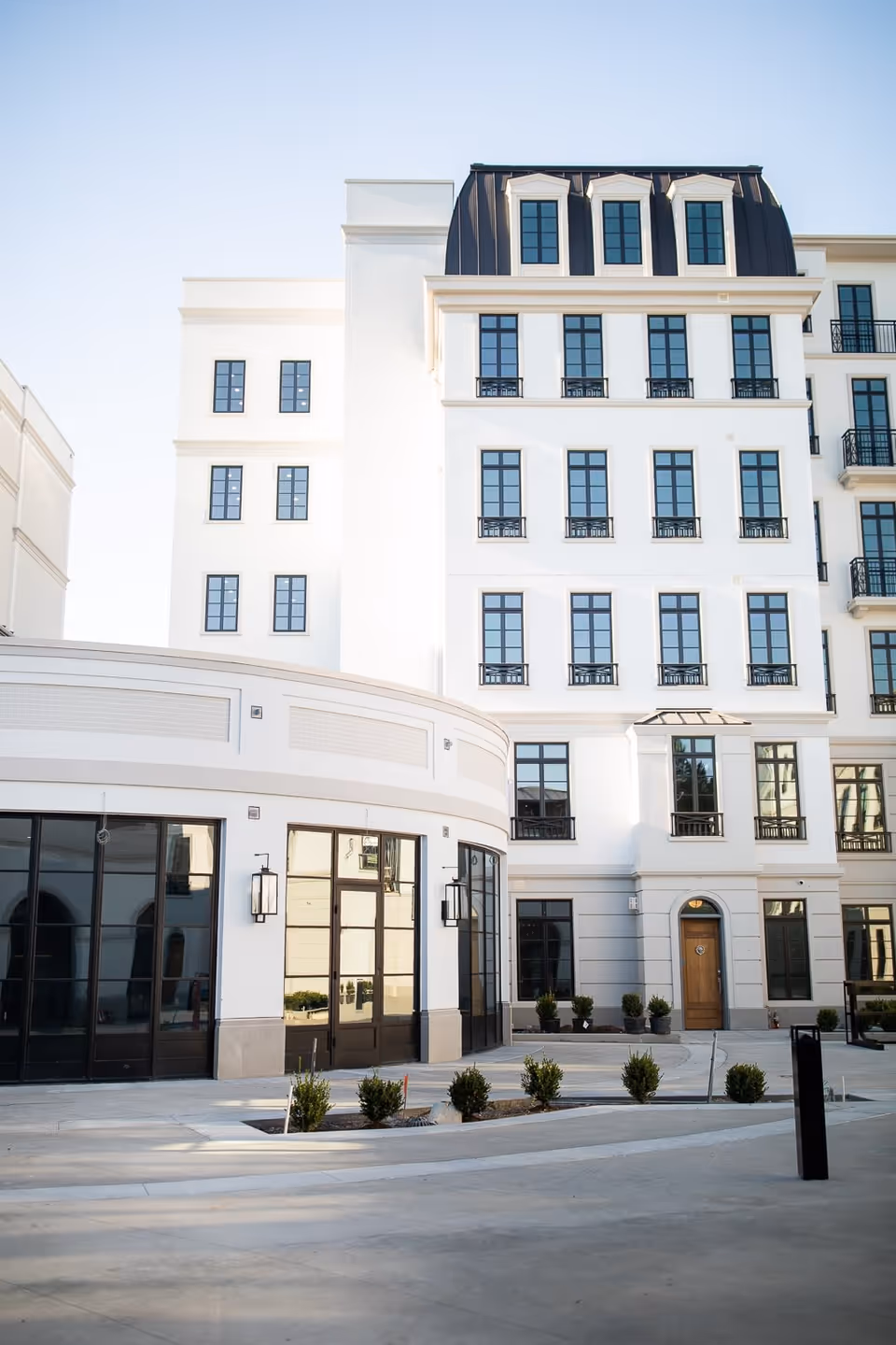 Exterior view of a modern multi-story white building with numerous windows and a curved entrance area with large glass doors. Small shrubs are planted along the paved area in front of the building under a clear sky.