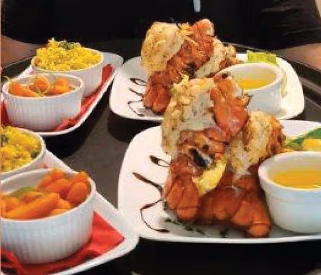 Close-up view of plates with lobster tails, dipping sauce, and side dishes including cooked carrots and yellow rice, served on a black table.
