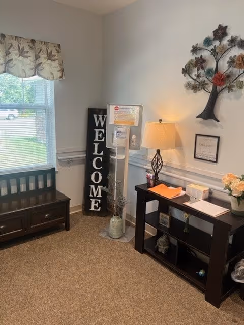 A cozy corner of a room with a window covered by a floral valance, a dark wooden bench with storage drawers, a tall vertical sign that says 'WELCOME', a hand sanitizer stand, a dark wooden console table with a lamp, some papers, a small plant, and decorative items. On the wall above the table is a metal tree wall art and a framed quote.