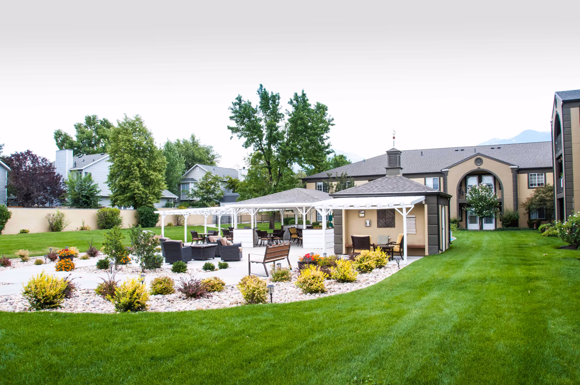 Outdoor seating area with multiple covered gazebos, chairs, and tables surrounded by well-maintained green grass, shrubs, and trees. Residential buildings are visible in the background under a clear sky.
