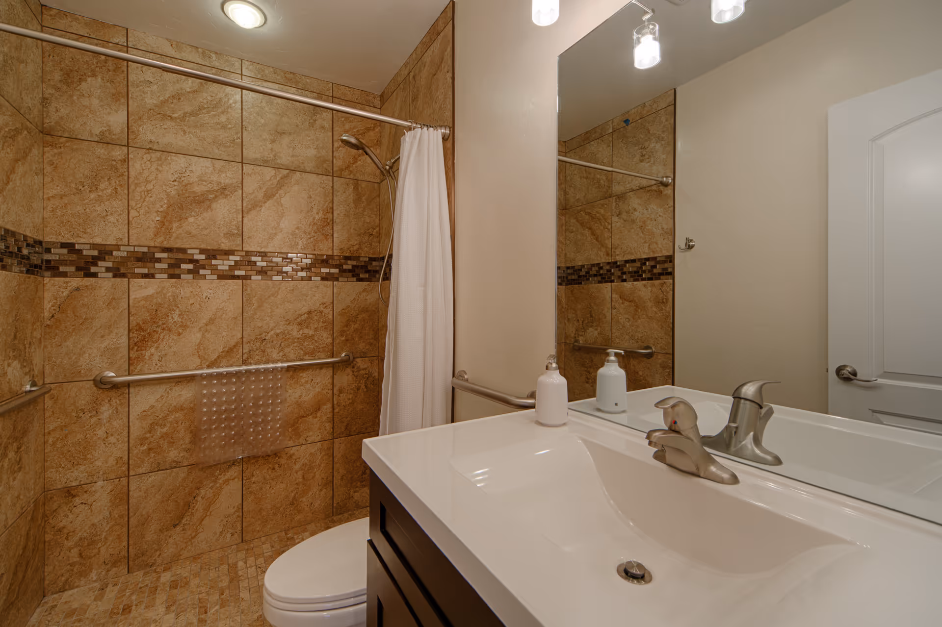 A bathroom with beige and brown tiled walls and floor, a white toilet, a white sink with a silver faucet, a large mirror above the sink, a white soap dispenser, a shower area with a white curtain, and metal grab bars on the walls.
