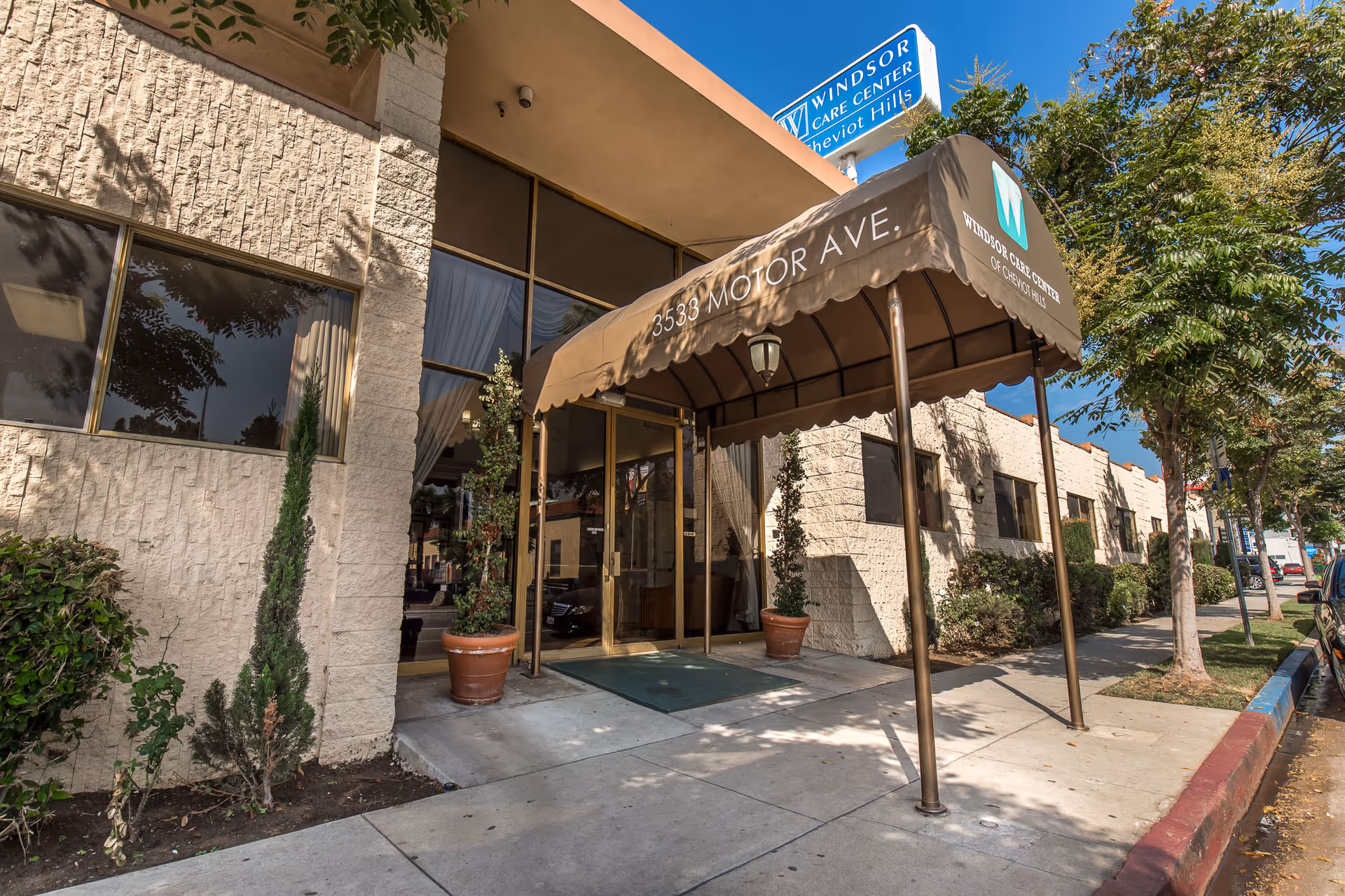 Entrance of Windsor Care Center of Cheviot Hills with a brown canopy displaying the address 3533 Motor Ave. The building has beige textured walls, large windows, potted plants, and a sidewalk with trees and parked cars along the street.