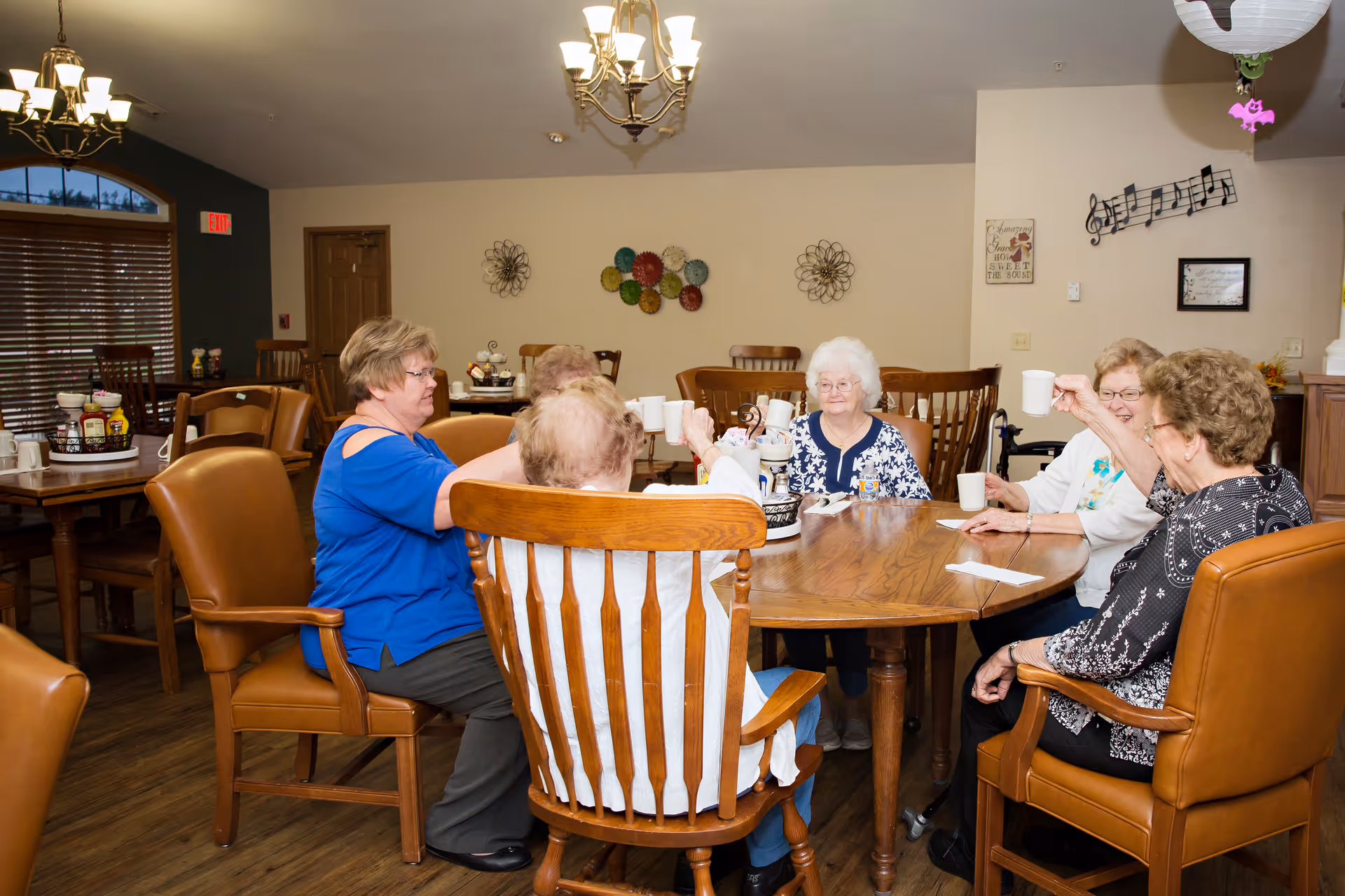 A group of elderly women sitting around a wooden dining table in a well-lit room, raising white mugs in a toast. The room has wooden chairs, wall decorations including musical notes, and chandeliers hanging from the ceiling.