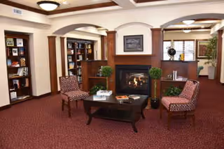 A cozy living room area with a fireplace in the center, flanked by two wooden columns and built-in bookshelves. There are two patterned armchairs facing a dark wooden coffee table with books and decorative items on it. The room has warm lighting, a red carpet, and cream-colored walls with crown molding.