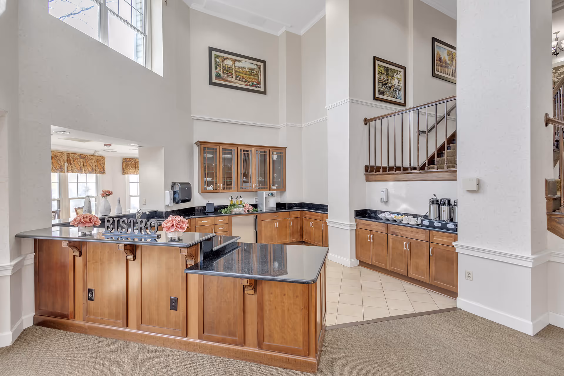 Interior view of a senior living facility's bistro area with wooden cabinets, black countertops, a coffee station, and decorative flowers. The space features high ceilings, framed artwork on the walls, and a staircase with a wooden railing.