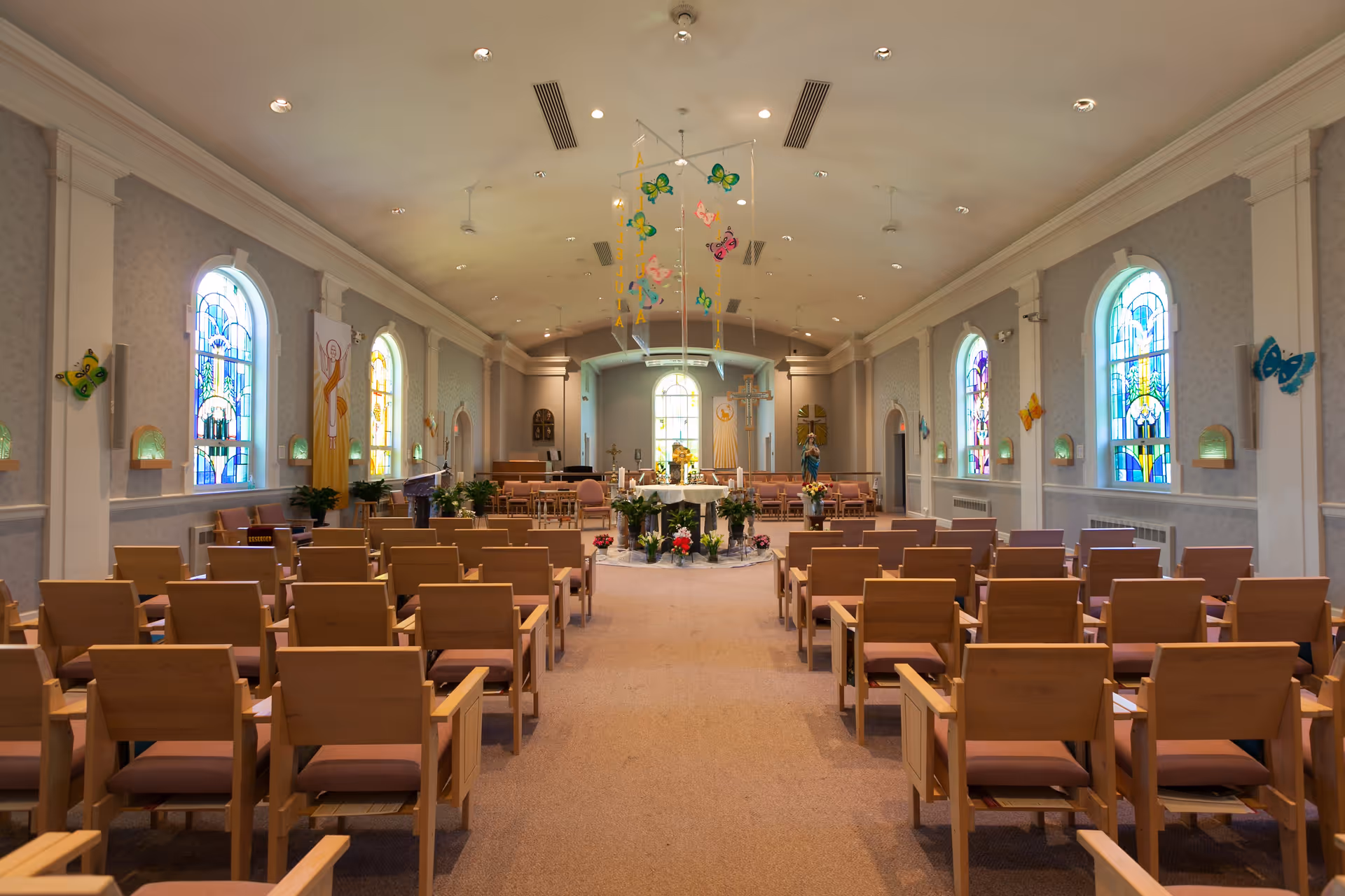 Interior view of a chapel with rows of wooden chairs facing an altar decorated with flowers. The chapel has arched stained glass windows on both sides and colorful butterfly decorations hanging from the ceiling and walls.