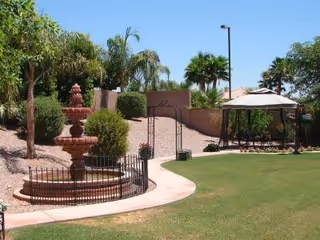 Outdoor garden area with a multi-tiered water fountain surrounded by a small black fence, palm trees, bushes, a paved walkway, and a gazebo with seating under a beige canopy.