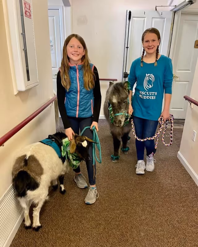 Two young girls standing in a hallway inside a building, each holding a leash. One girl is holding a goat with a green bandana, and the other girl is holding a small pony wearing green accessories. The girls are smiling and the hallway has beige walls and carpeted floor.
