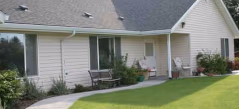 Single-story building with light siding, curved walkway, patio chairs, and manicured lawn outside.