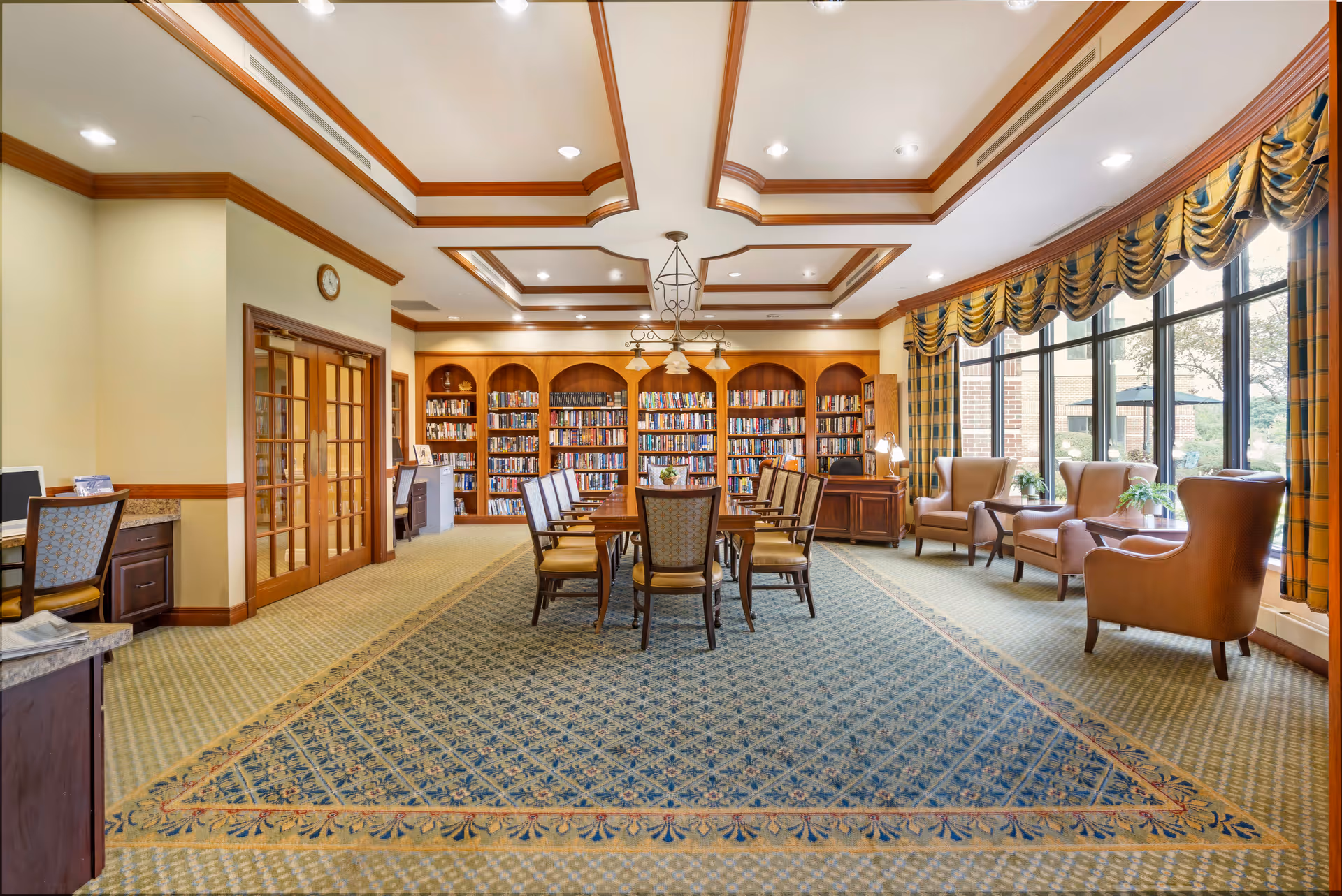 A spacious and well-lit library or reading room with a large wooden table surrounded by chairs in the center. The back wall features built-in wooden bookshelves filled with books. To the right, there are several comfortable armchairs next to large windows with yellow and blue plaid curtains, allowing natural light to fill the room. The ceiling has decorative wooden beams and recessed lighting.