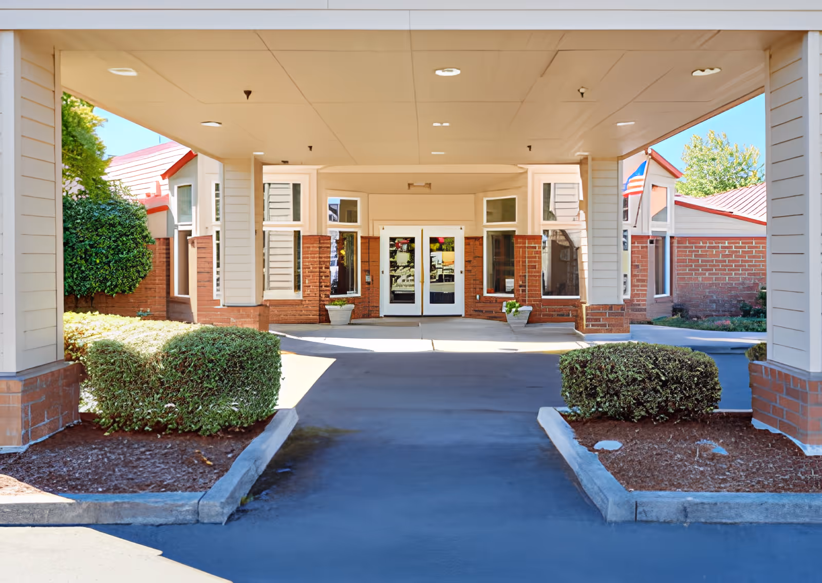 Covered entrance driveway leading to double glass doors of a brick and siding building with neatly trimmed bushes on either side and an American flag visible on the right side.