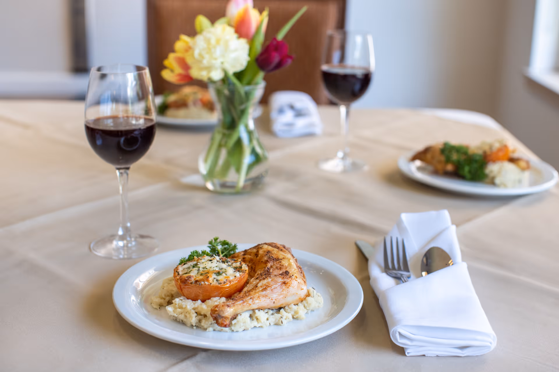 A dining table set with plates of food including a roasted chicken leg on a bed of mashed potatoes and a stuffed tomato, two glasses of red wine, a folded white napkin with silverware, and a vase with colorful flowers in the background.