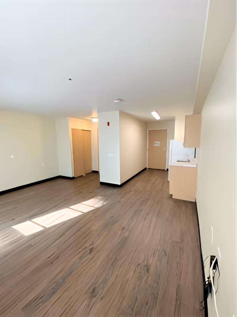 Empty senior living studio interior with wood-look flooring, a small kitchenette on the right, and a hallway/closet and entry door at the back.