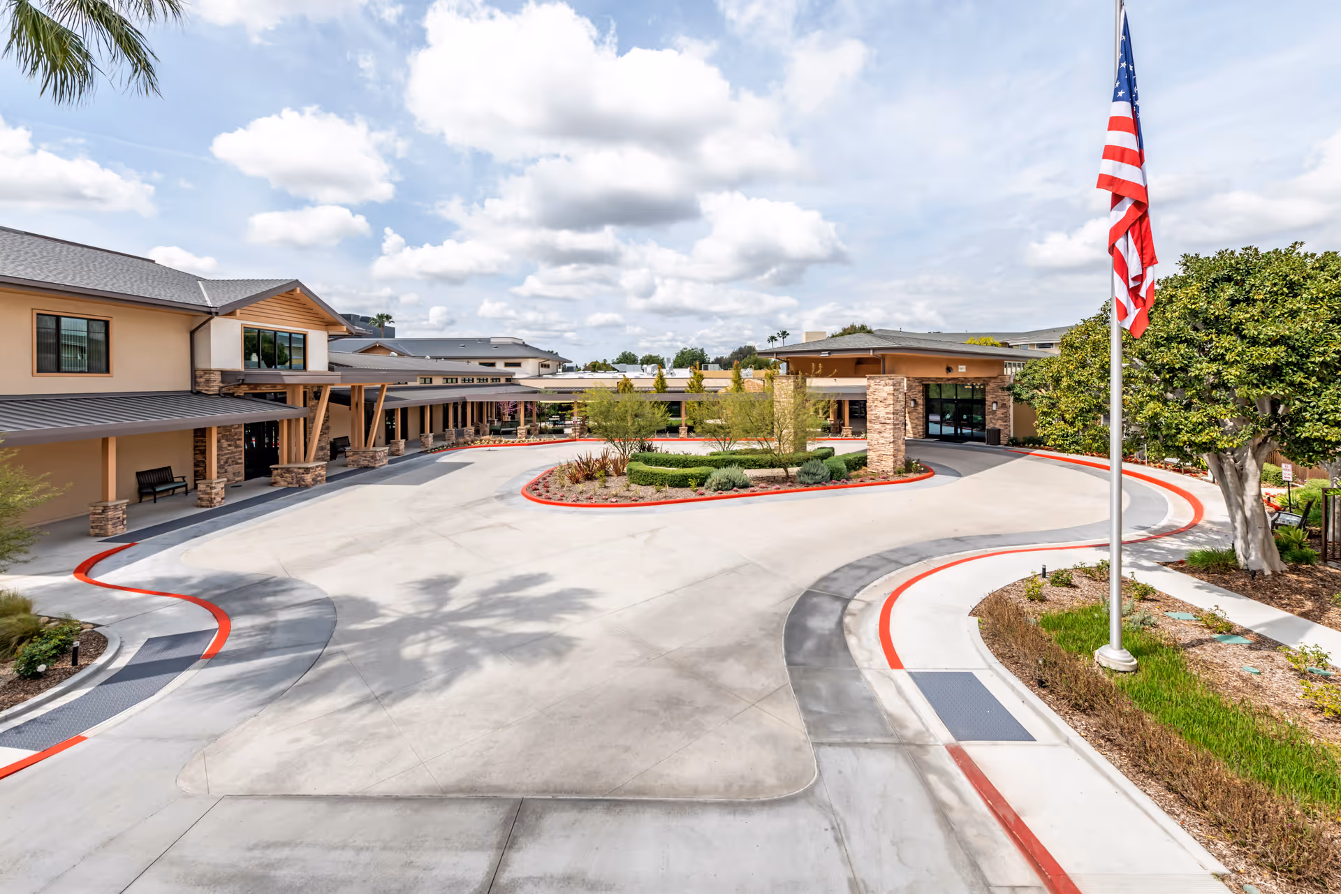 Exterior view of Emerald Court senior living facility showing a curved driveway with red curbs, landscaped greenery in the center, a flagpole with an American flag, and a building with stone and wood accents under a partly cloudy sky.