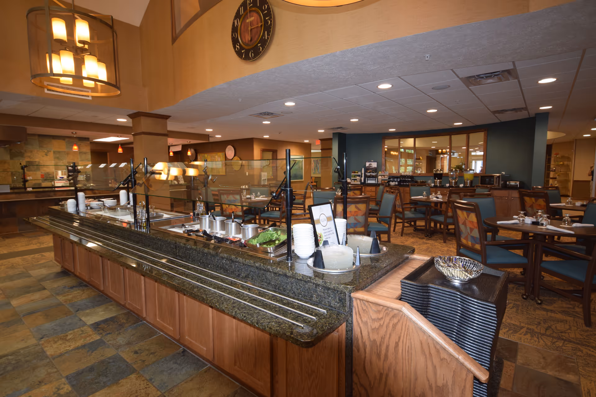 Interior view of a dining area in a retirement community featuring a buffet counter with food warmers, plates, and utensils. The room has multiple tables and chairs arranged for dining, warm lighting fixtures, and a large clock on the wall.