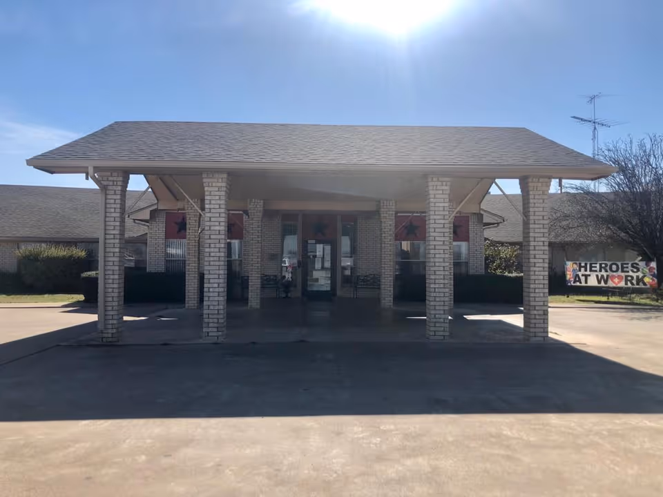 Entrance canopy of a brick senior living facility with multiple columns and a 'HEROES AT WORK' sign visible to the right.