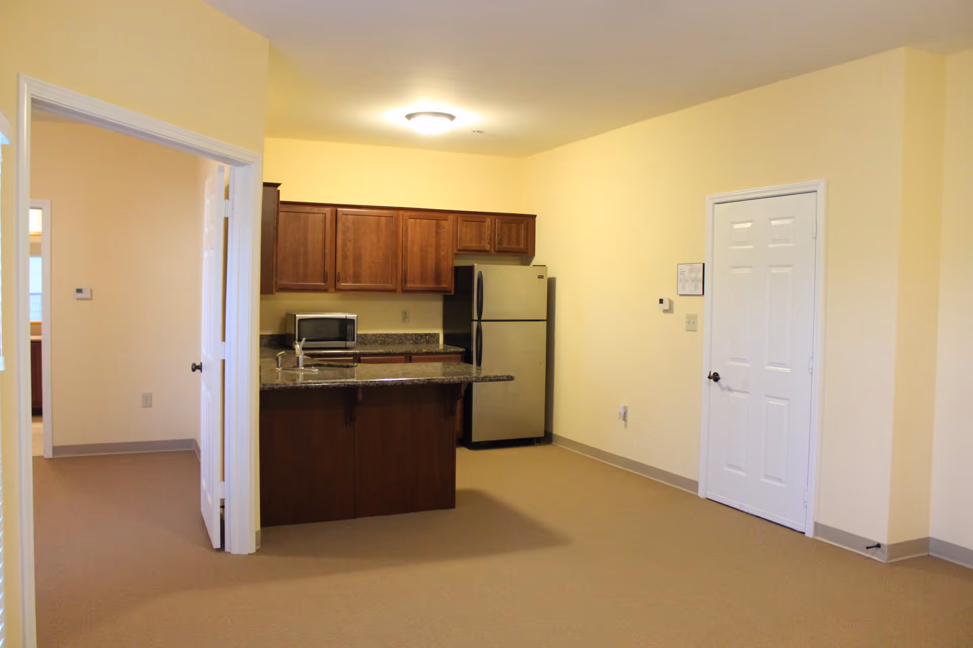 Interior view of a senior living facility apartment showing a small kitchen area with wooden cabinets, a granite countertop island, a microwave, and a stainless steel refrigerator. There is an open doorway leading to another room and a closed white door on the right wall. The walls are painted light yellow and the floor is carpeted.