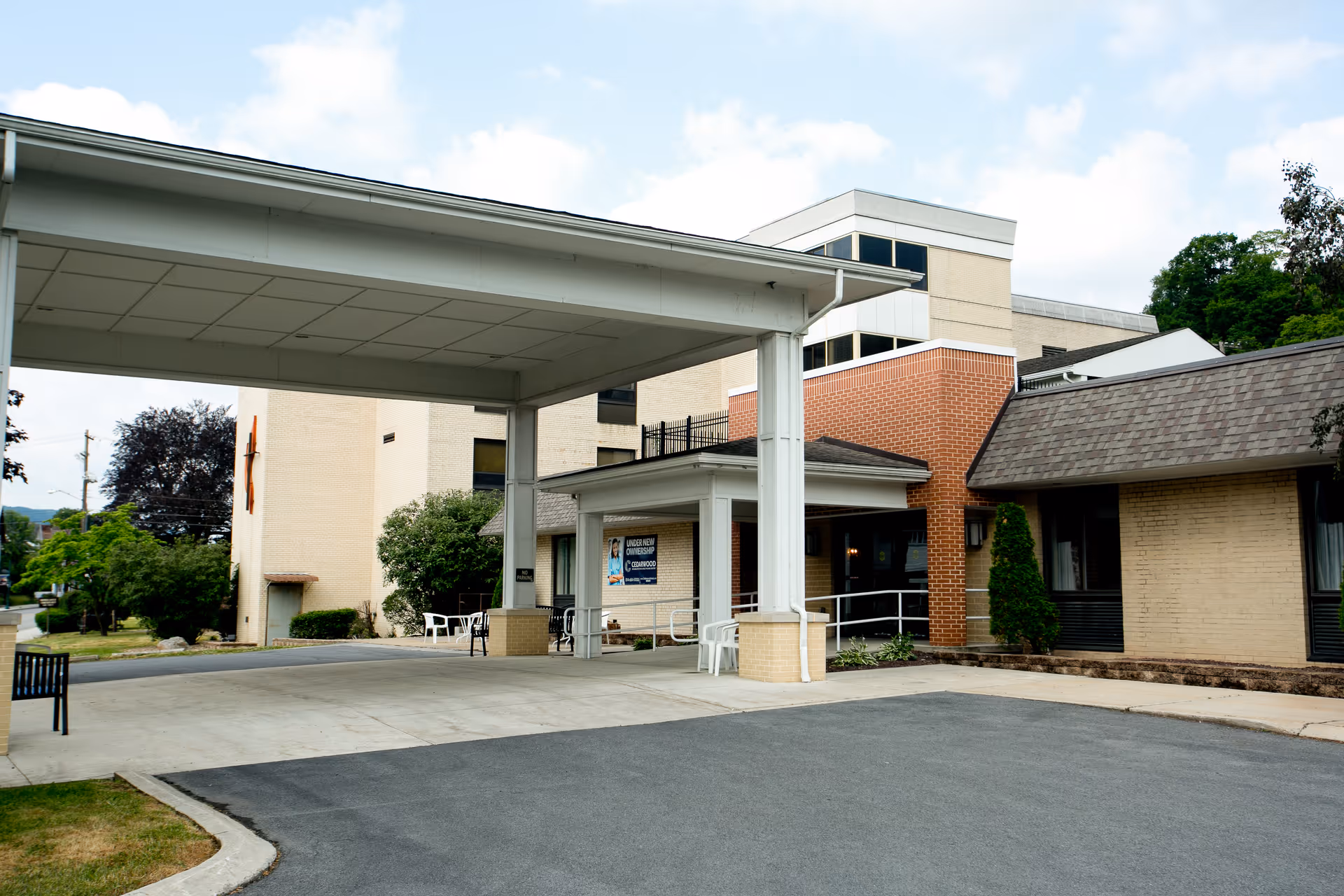 Exterior view of Cedarwood Rehabilitation and Healthcare Center showing the entrance with a covered drop-off area, brick and beige building facade, some greenery, and a clear sky.