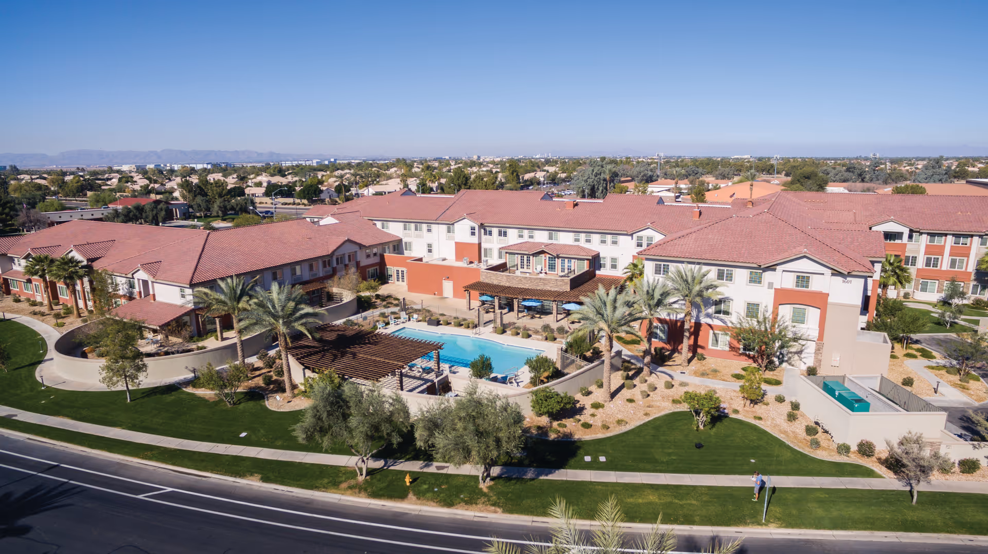 Aerial view of the Gardens at Ocotillo senior living community with red-tile roof buildings, a central swimming pool, pergolas, palm trees and landscaped grounds.