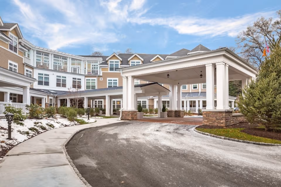 Exterior view of Sunrise of Fairfield senior living facility showing a large covered entrance with white columns and stone bases, a curved driveway, landscaped areas with some snow on the ground, and a multi-story building with numerous windows under a partly cloudy sky.