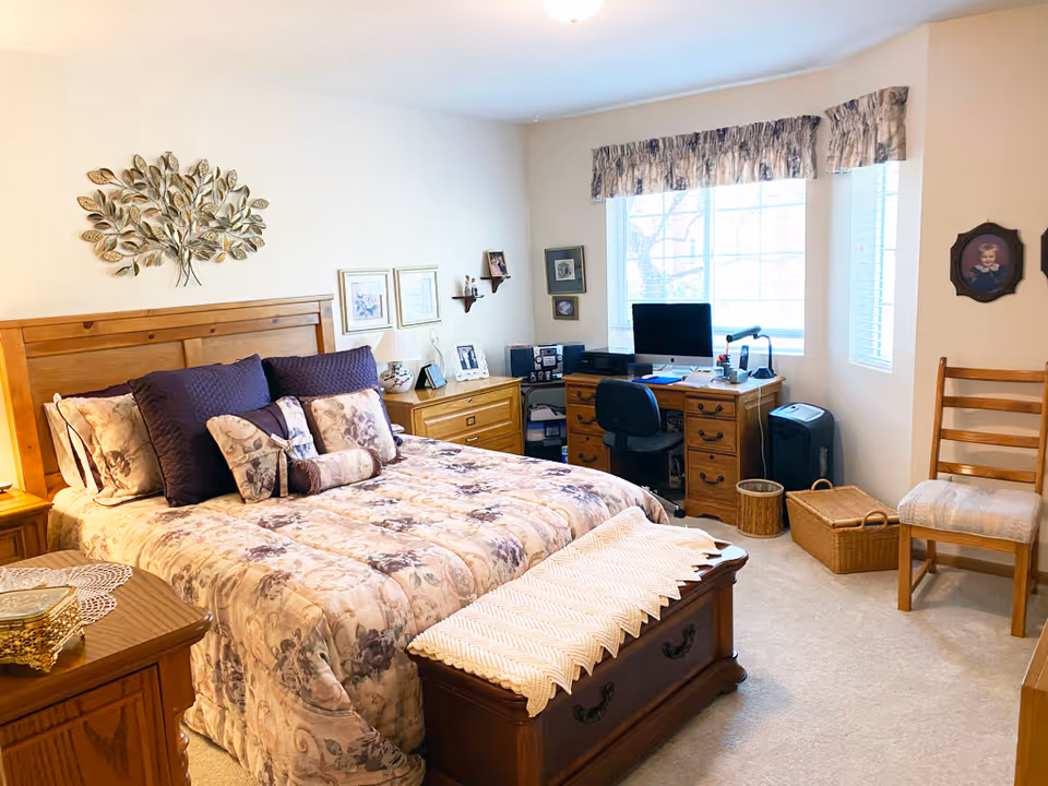 Well-lit bedroom with a floral comforter on a wooden bed, matching wooden furniture, a desk with a computer by a large window, and seating.